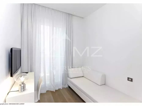 Minimalist white room with a wall-mounted TV, a white desk with a silver lamp, sheer curtains, and a white sofa along the right wall.