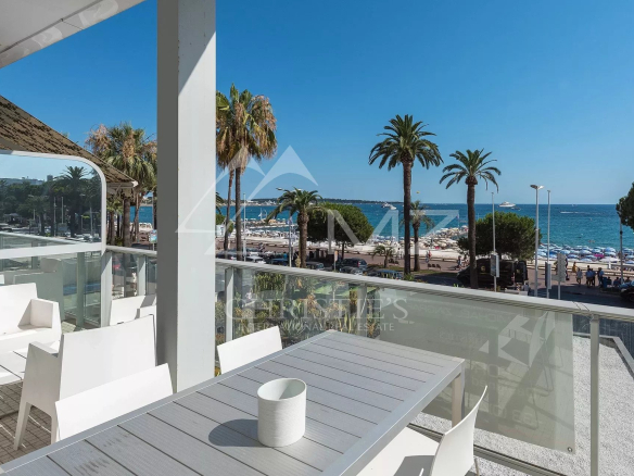 Outdoor white patio seating facing a sunny beach and blue sea, with palm trees and a glass railing nearby.