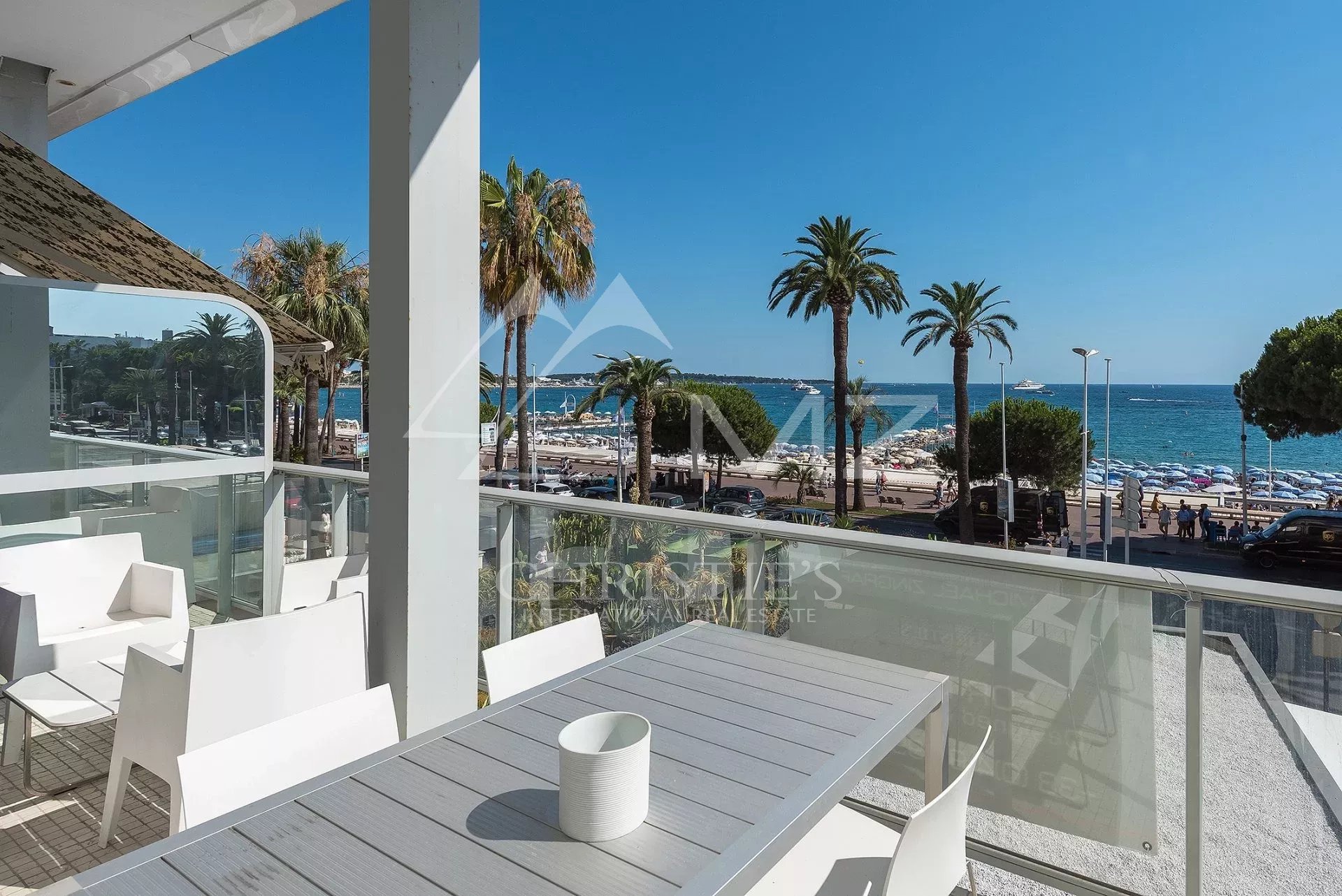 Outdoor white patio seating facing a sunny beach and blue sea, with palm trees and a glass railing nearby.