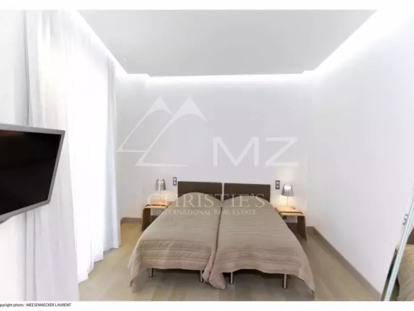 Twin-bed hotel room with beige bedspreads, two nightstands and lamps, a wall-mounted TV on the left, white walls and sheer curtains.