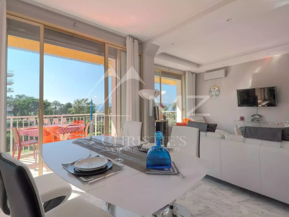 Open-plan living and dining area with white furniture, marble floor, and a sliding glass door to a sunlit balcony with orange outdoor seating and palm trees outside.