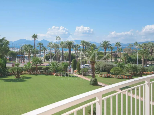View from a balcony of a coastal garden: a green lawn, palm trees, and flowering beds with a marina and mountains in the distance under a blue sky.
