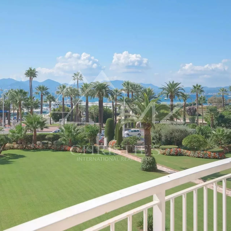 View from a balcony of a coastal garden: a green lawn, palm trees, and flowering beds with a marina and mountains in the distance under a blue sky.