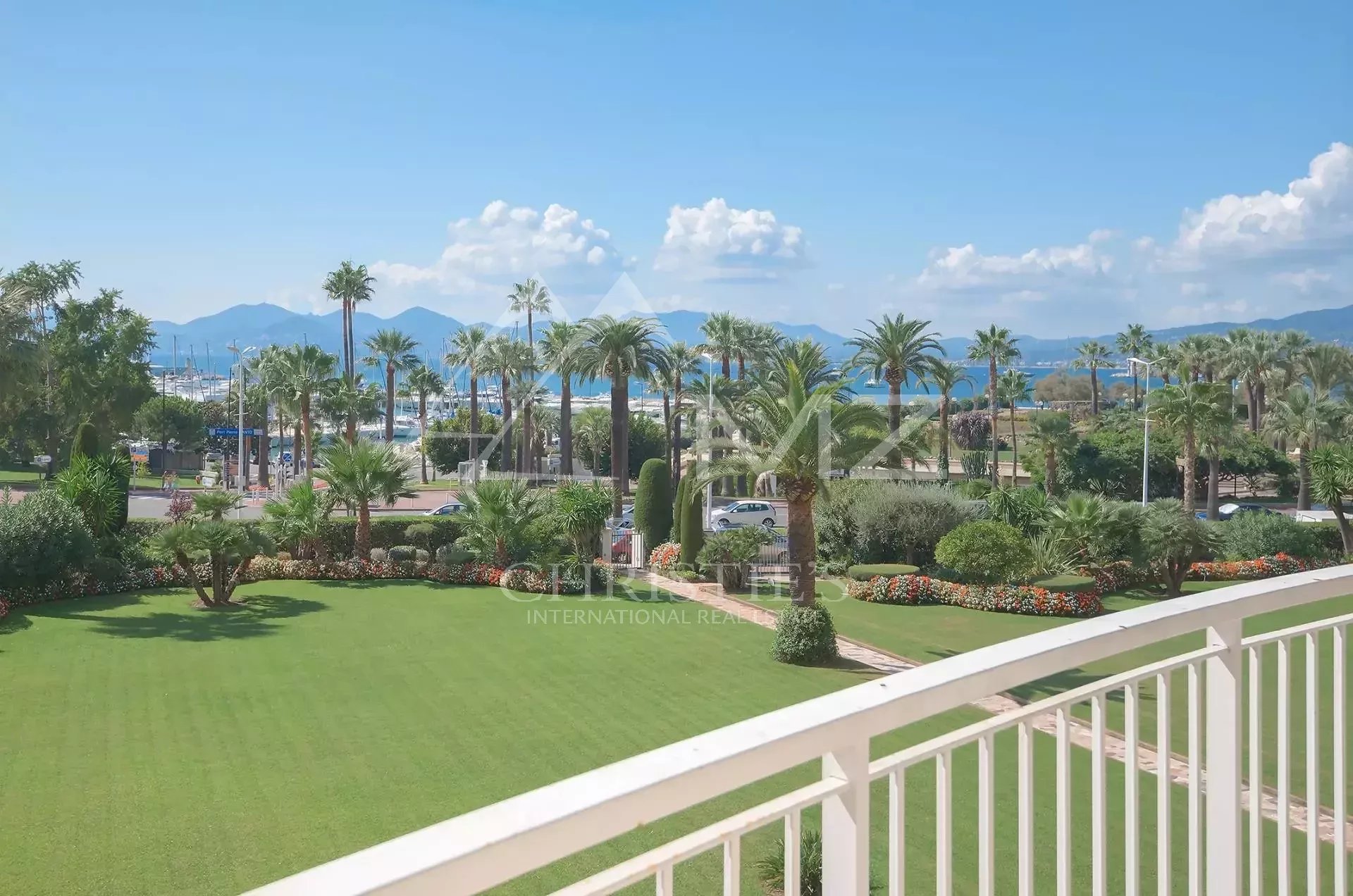 View from a balcony of a coastal garden: a green lawn, palm trees, and flowering beds with a marina and mountains in the distance under a blue sky.