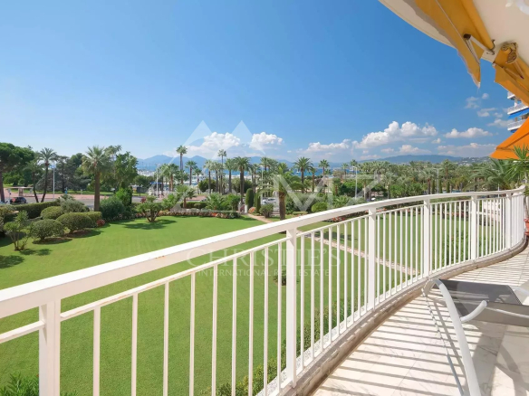 Balcony railing overlooking a green lawn with palm trees and a distant blue sea under a clear sky, with lounge chairs in the foreground to the right.