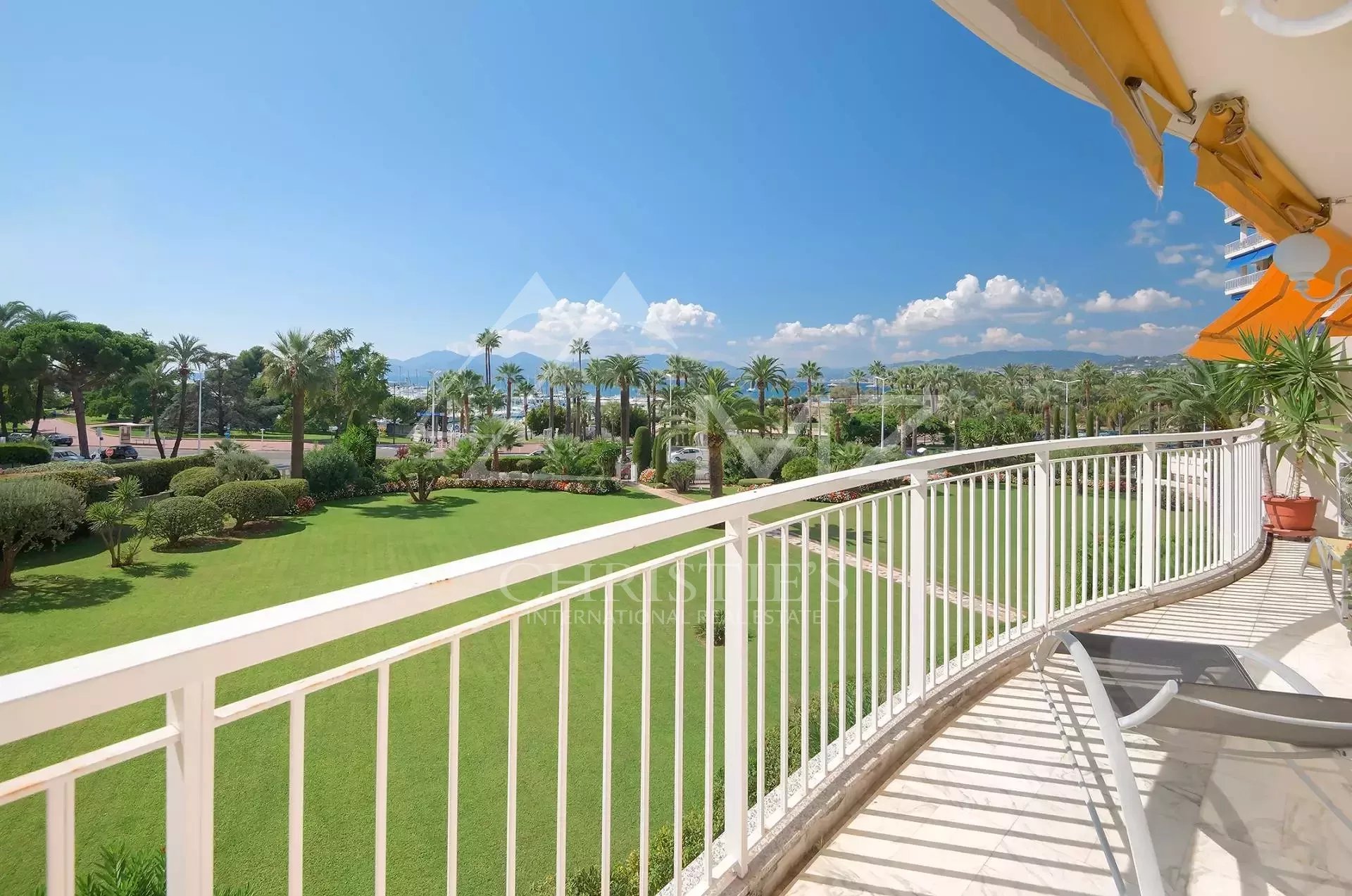 Balcony railing overlooking a green lawn with palm trees and a distant blue sea under a clear sky, with lounge chairs in the foreground to the right.