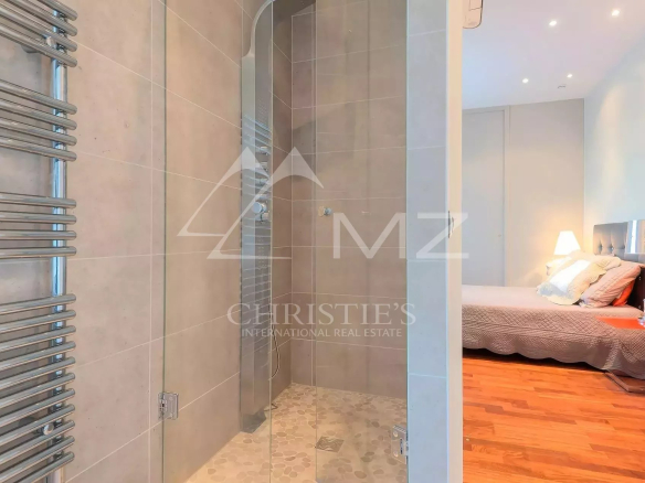 Glass-enclosed shower with pebble floor adjacent to a bedroom, chrome towel radiator on the left and a warm-lit bed area beyond.