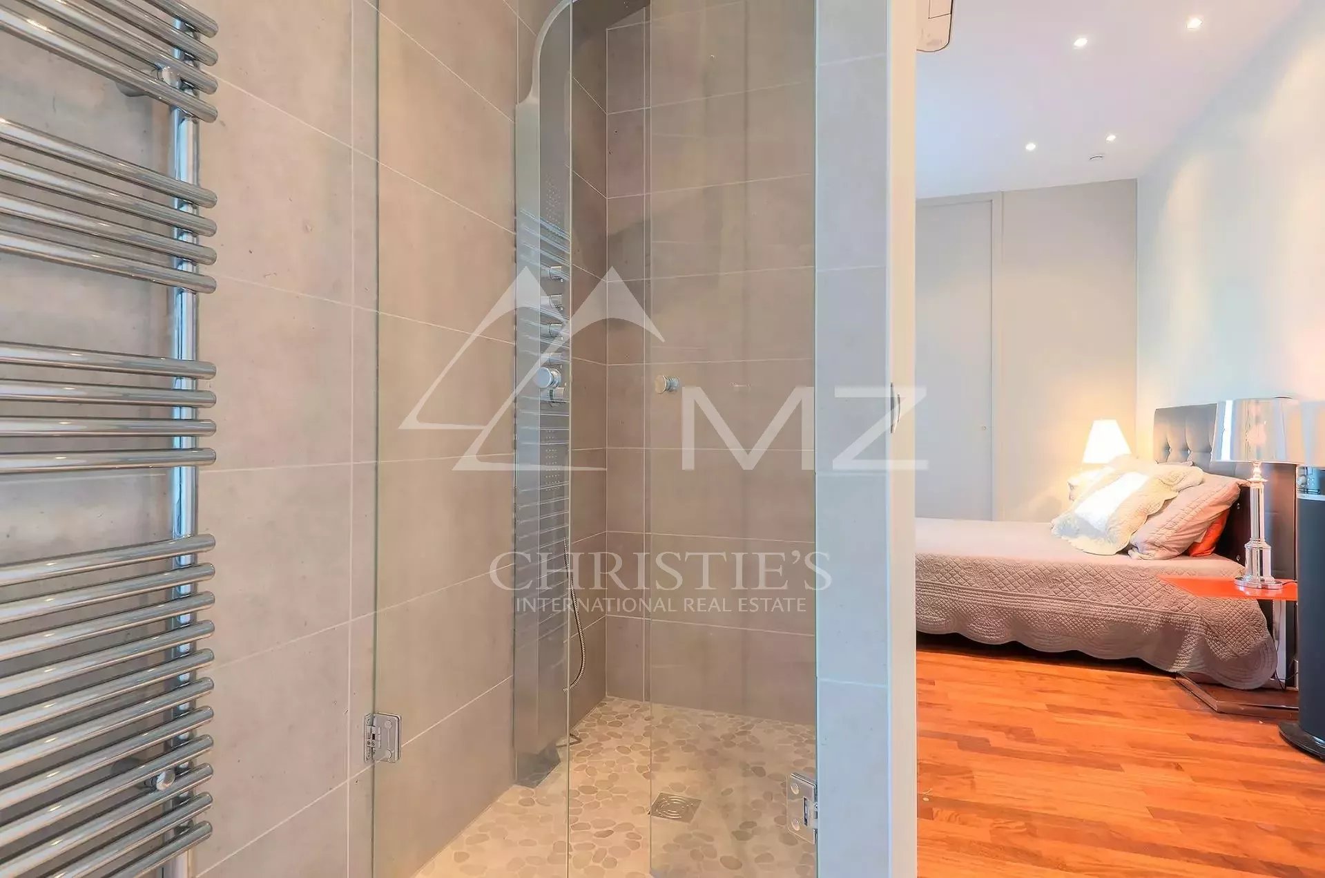 Glass-enclosed shower with pebble floor adjacent to a bedroom, chrome towel radiator on the left and a warm-lit bed area beyond.
