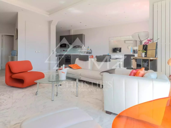 Open-plan living room with white sofas and a bright orange lounge chair, glass coffee table, and a modern kitchen in the background.