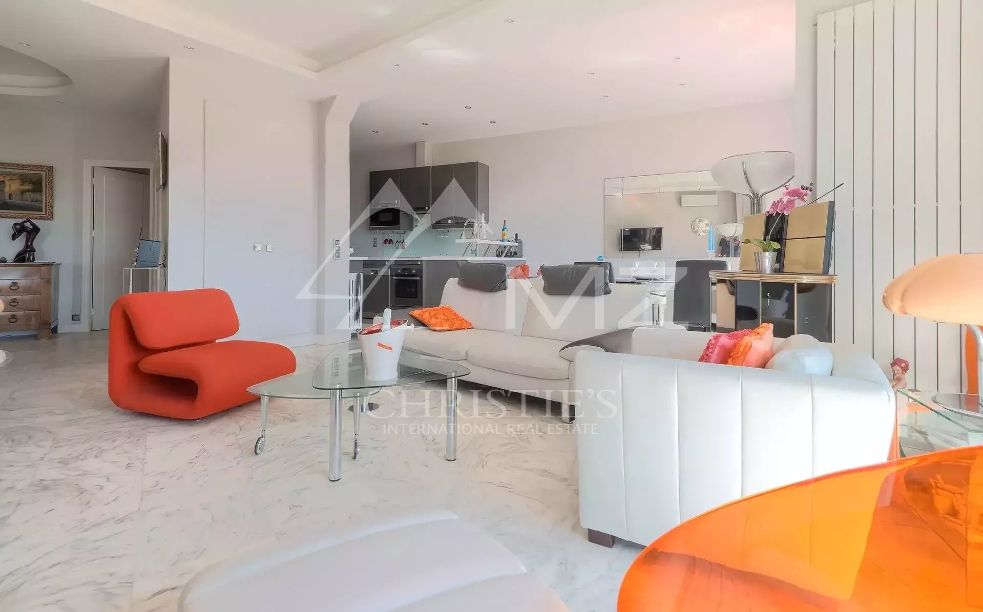 Open-plan living room with white sofas and a bright orange lounge chair, glass coffee table, and a modern kitchen in the background.