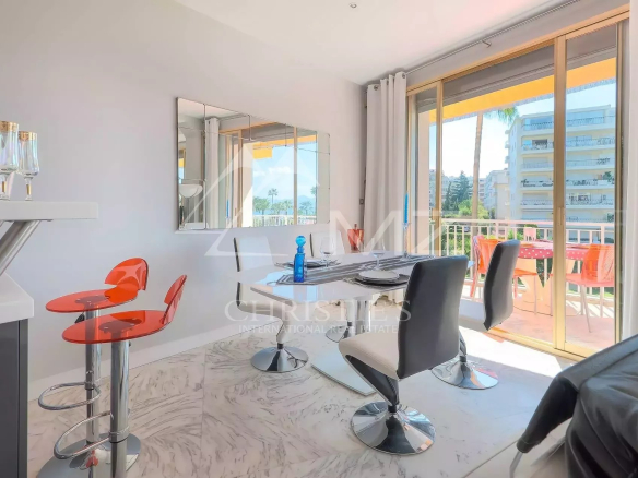 Bright dining area with a marble floor, a white table set for four, and orange bar stools near a champagne bottle on a small counter. Sliding glass doors open to a sunny balcony with orange chairs outside.