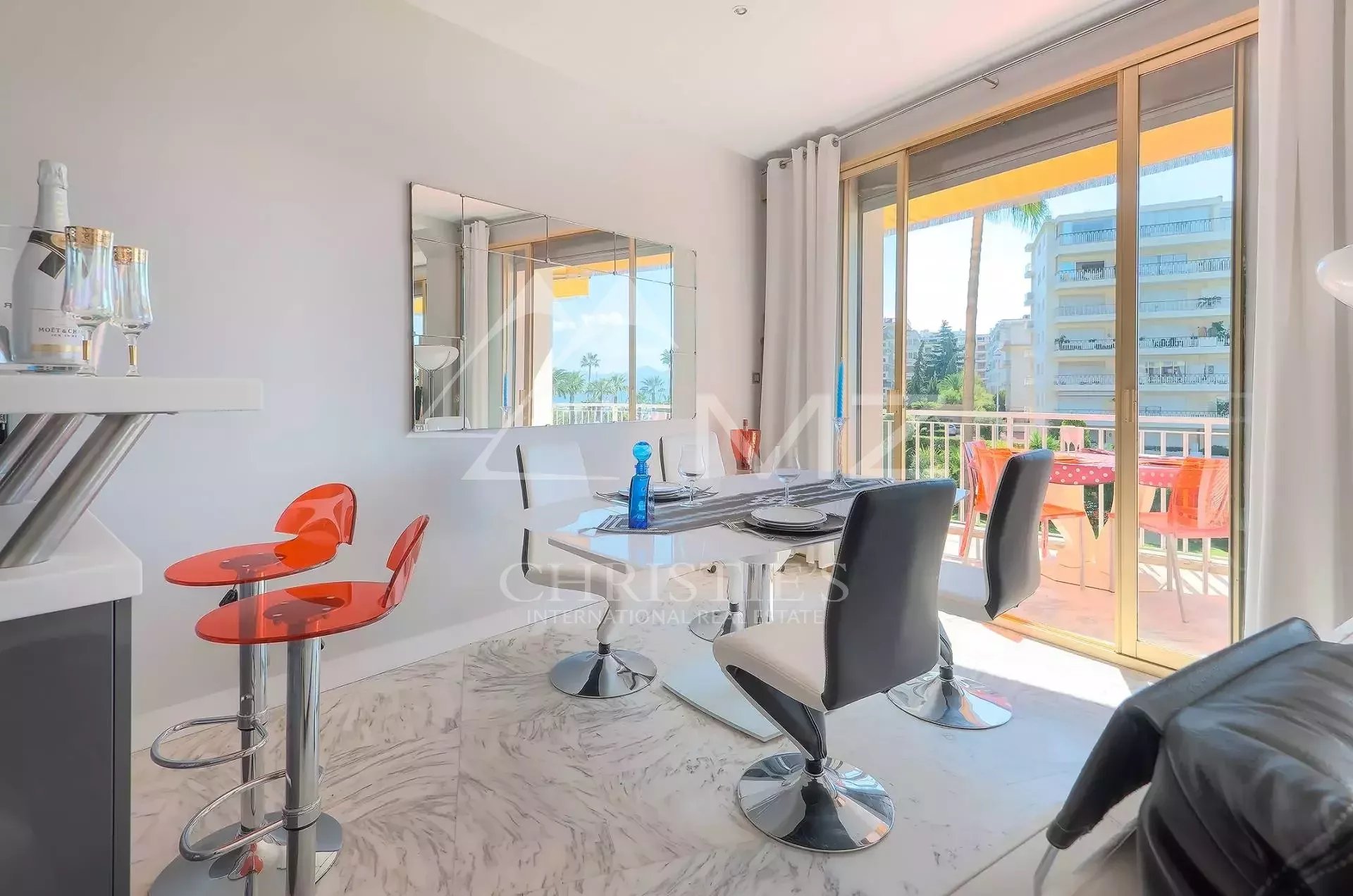 Bright dining area with a marble floor, a white table set for four, and orange bar stools near a champagne bottle on a small counter. Sliding glass doors open to a sunny balcony with orange chairs outside.