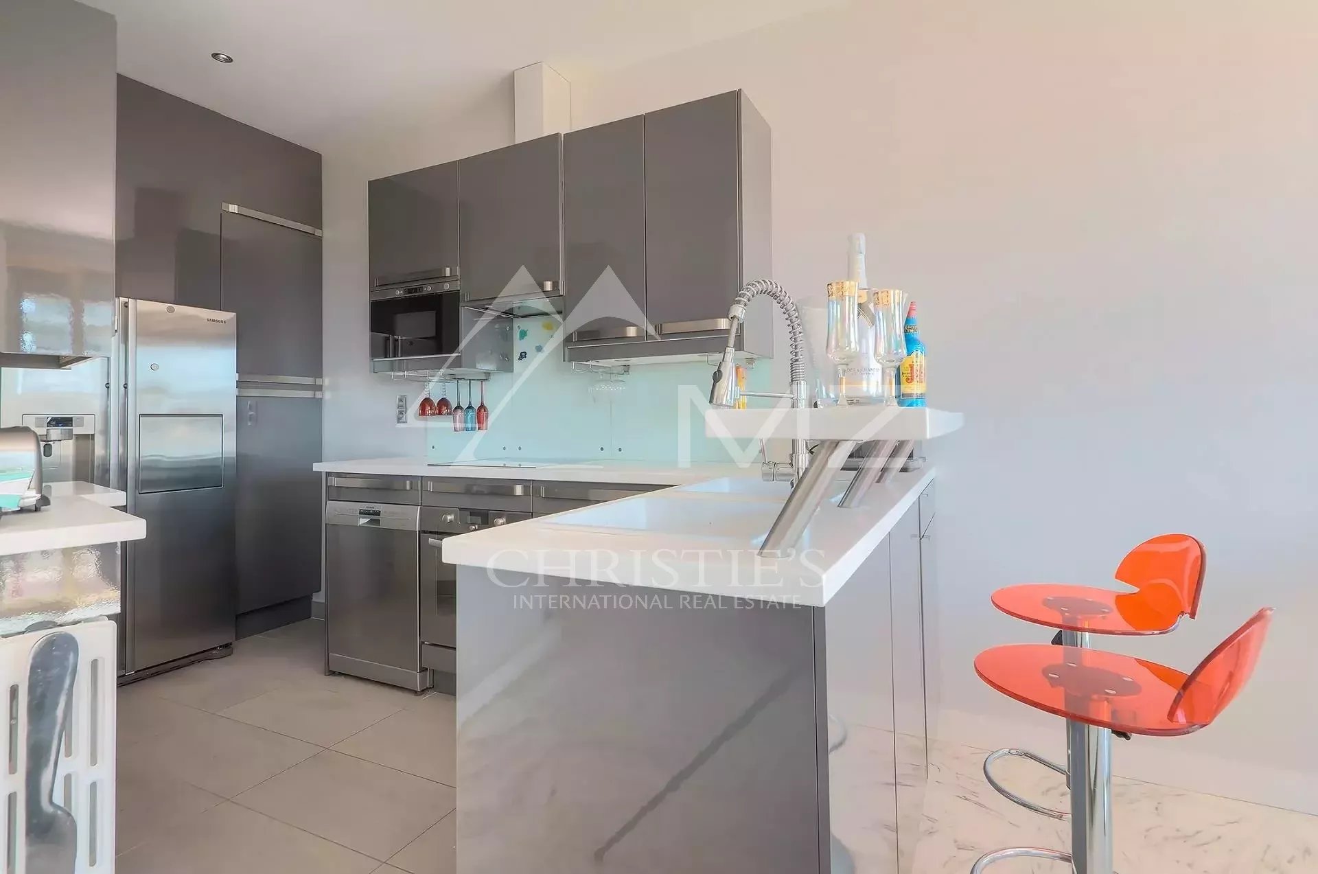 Modern kitchen with gray high-gloss cabinets, stainless steel fridge and built-in oven/m microwave, and a white island with a sink.