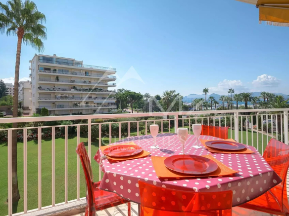 Balcony table set for four with pink polka-dot tablecloth, orange plates, and red chairs, overlooking a green lawn and palm trees toward the sea.