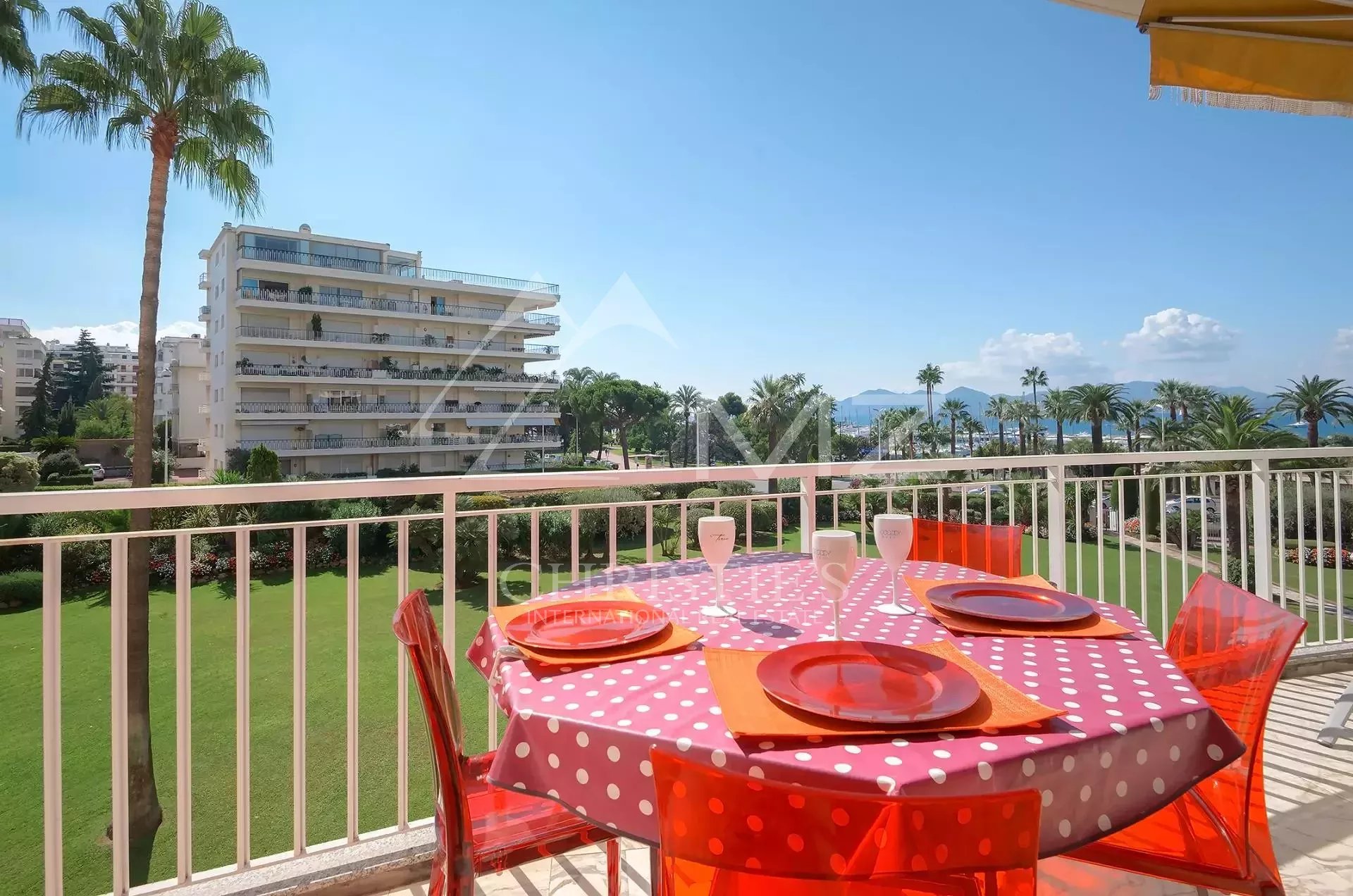 Balcony table set for four with pink polka-dot tablecloth, orange plates, and red chairs, overlooking a green lawn and palm trees toward the sea.