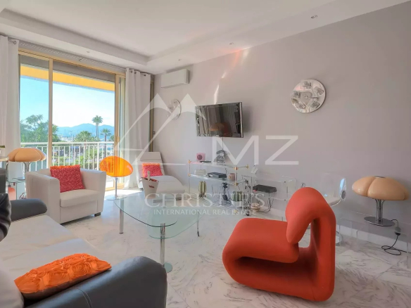 Contemporary living room with orange flamingo chair, white sofas, glass coffee table, and a wall-mounted TV; large balcony door shows palm trees outside.