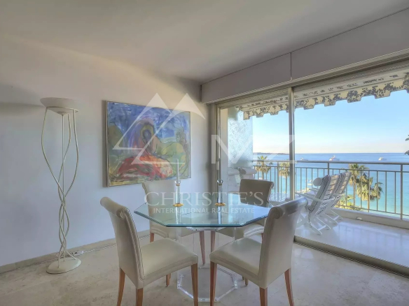 Bright dining area with a glass round table and four beige chairs, next to a sculptural floor lamp, overlooking a beachy balcony sea view.