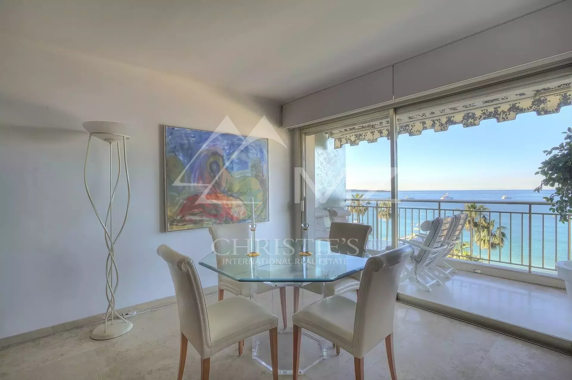 Bright dining area with a glass round table and four beige chairs, next to a sculptural floor lamp, overlooking a beachy balcony sea view.