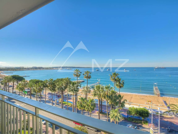 Balcony view of a palm-lined promenade and sandy beach with calm blue sea under a clear sky.