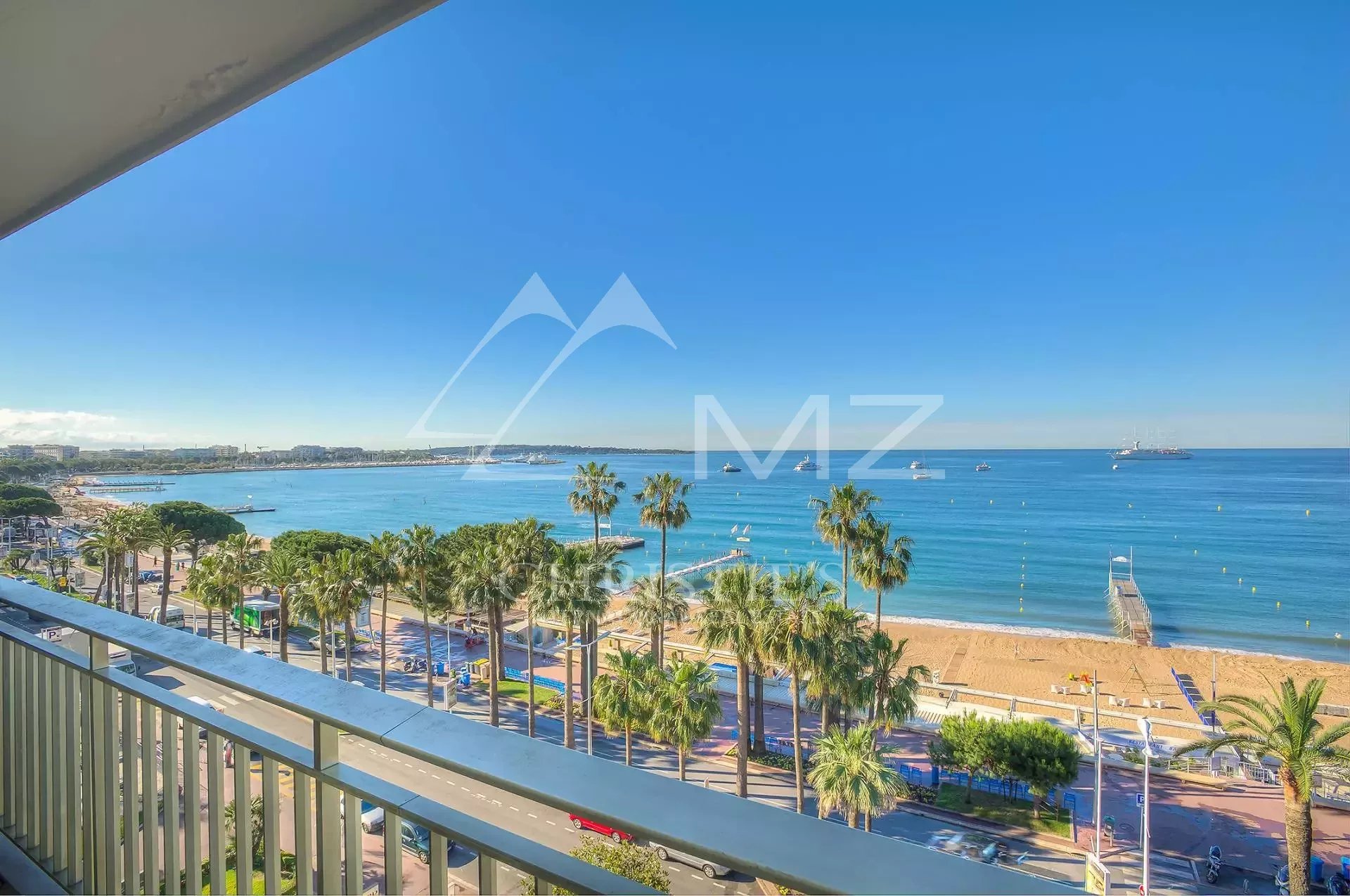 Balcony view of a palm-lined promenade and sandy beach with calm blue sea under a clear sky.
