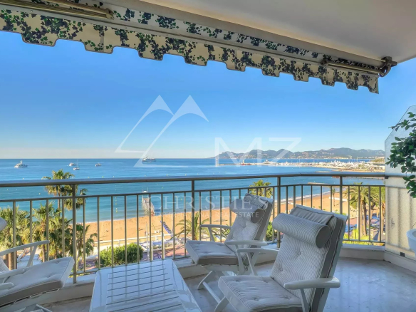 Balcony with white lounge chairs overlooking a sandy beach, palm trees, and a calm blue sea with boats and distant mountains.
