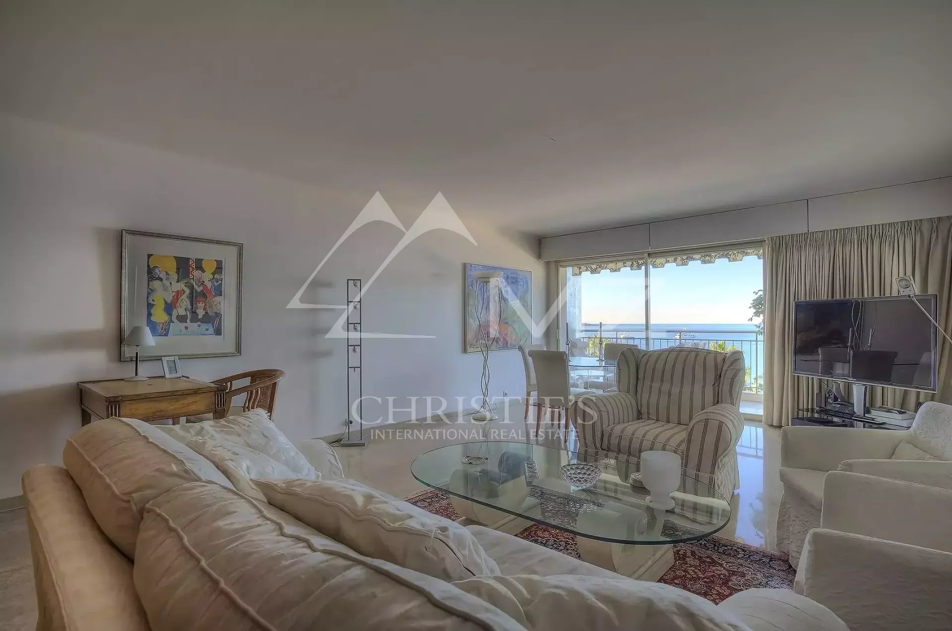 Bright living room with beige sofas, glass coffee table, and a balcony sea view through large sliding doors.