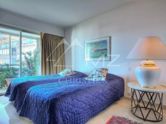 Hotel room with two twin beds in blue quilts, decorative pillows, a marble-topped side table, and a large lamp by the wall.