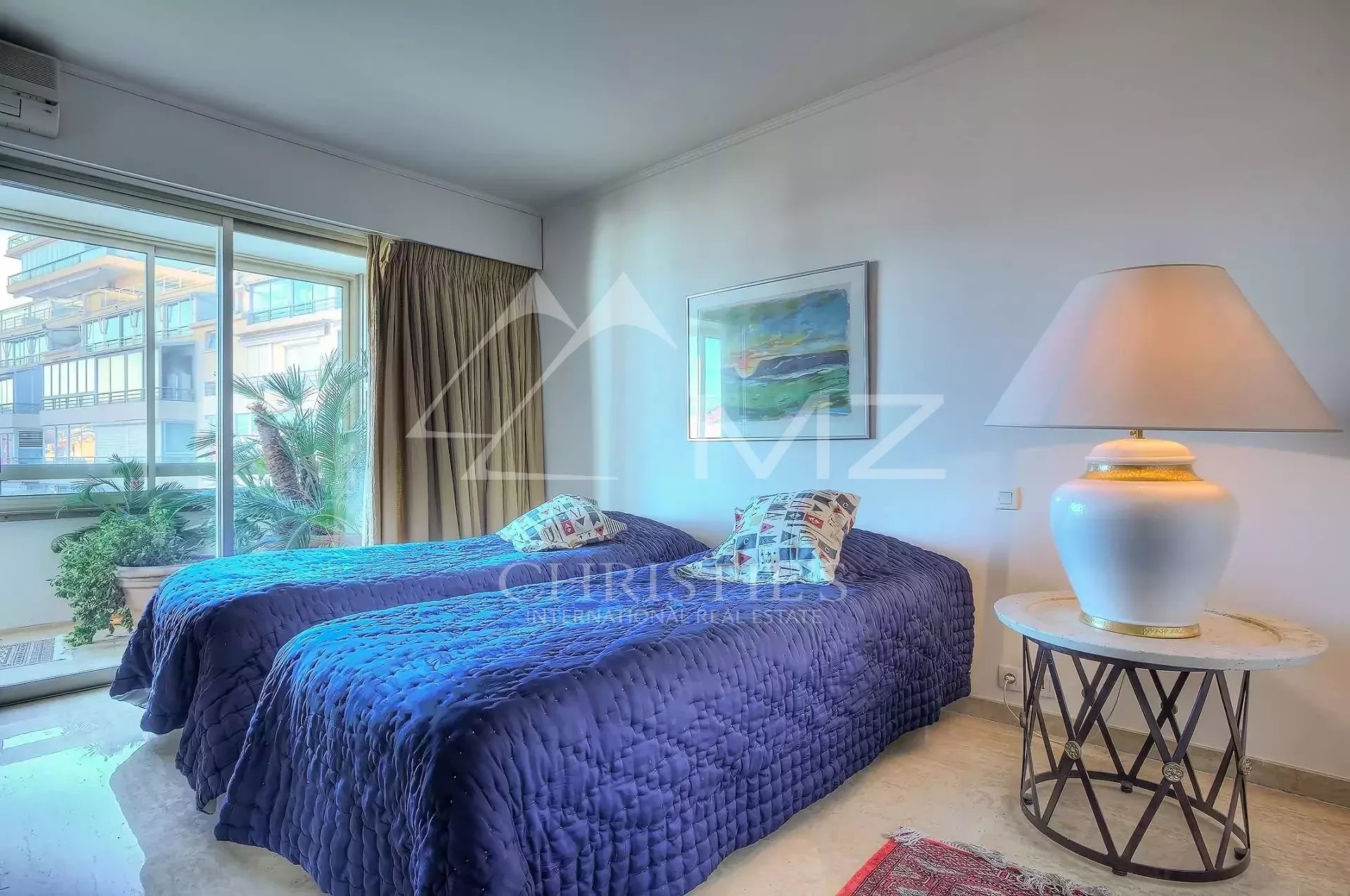 Hotel room with two twin beds in blue quilts, decorative pillows, a marble-topped side table, and a large lamp by the wall.