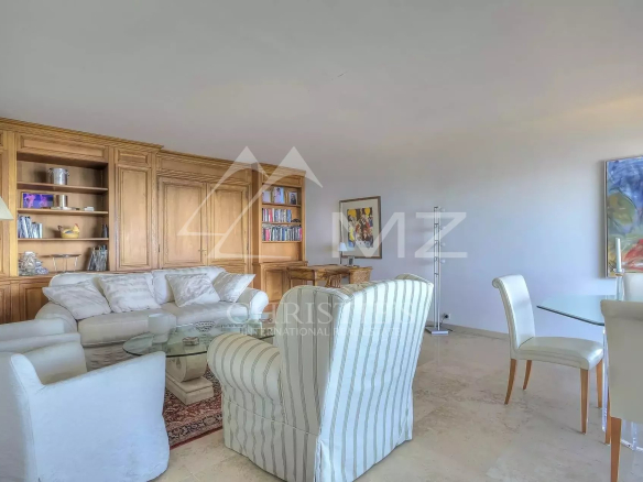 Bright living room with a cream sofa, striped armchairs, and a glass coffee table in front of wooden built-in shelves and cabinets. There is a marble floor and a floor lamp nearby.
