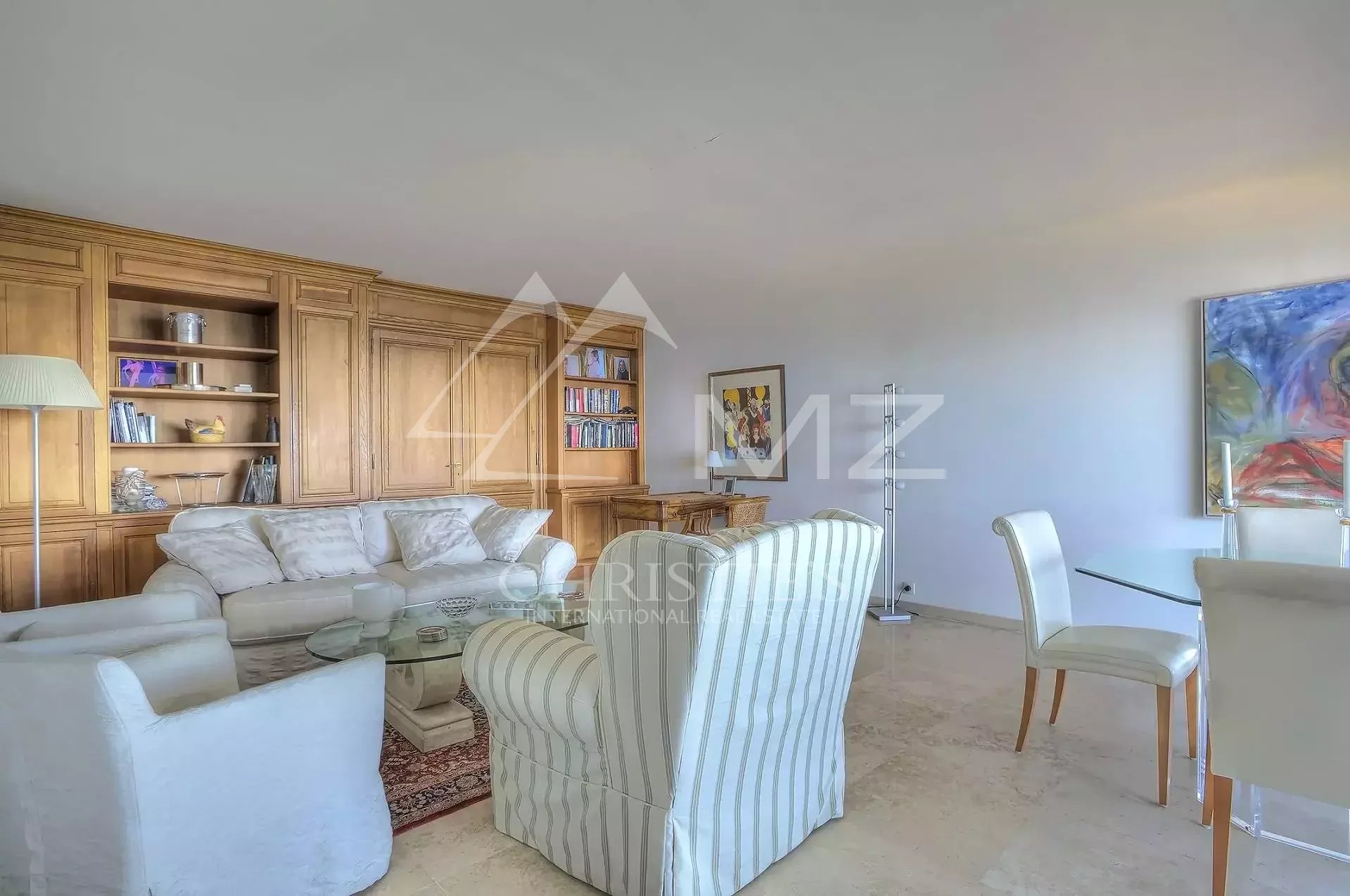 Bright living room with a cream sofa, striped armchairs, and a glass coffee table in front of wooden built-in shelves and cabinets. There is a marble floor and a floor lamp nearby.
