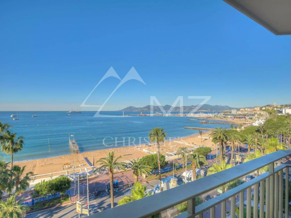 Sunny beach and calm blue sea viewed from a balcony, with palm trees and a promenade. (informative)"