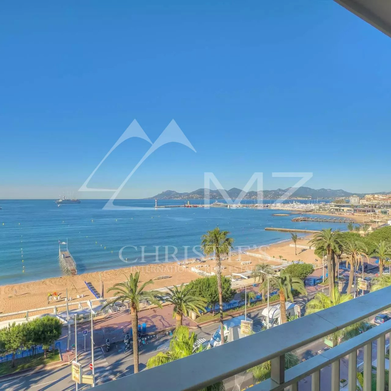 Sunny beach and calm blue sea viewed from a balcony, with palm trees and a promenade. (informative)"