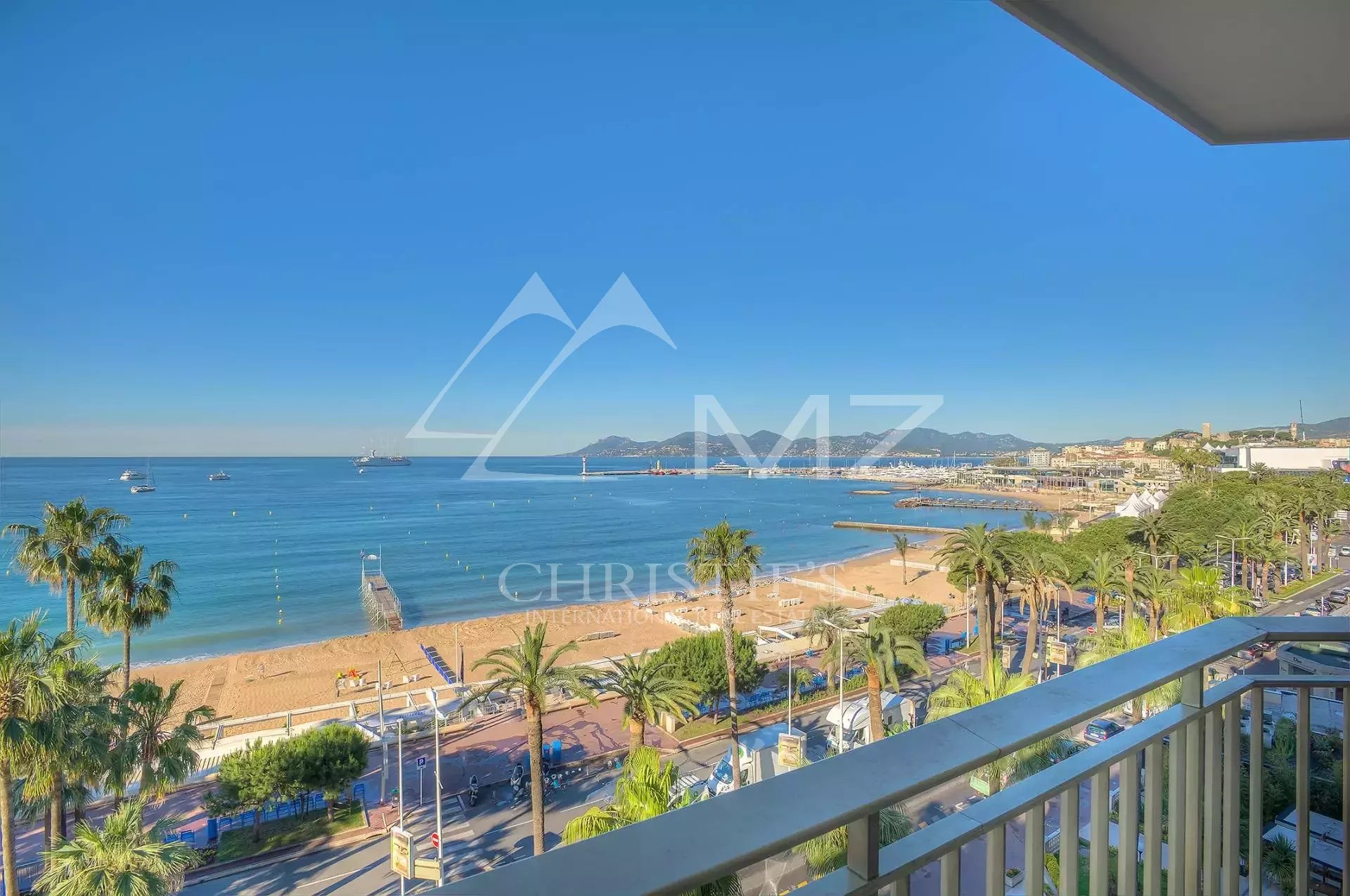 Sunny beach and calm blue sea viewed from a balcony, with palm trees and a promenade. (informative)"