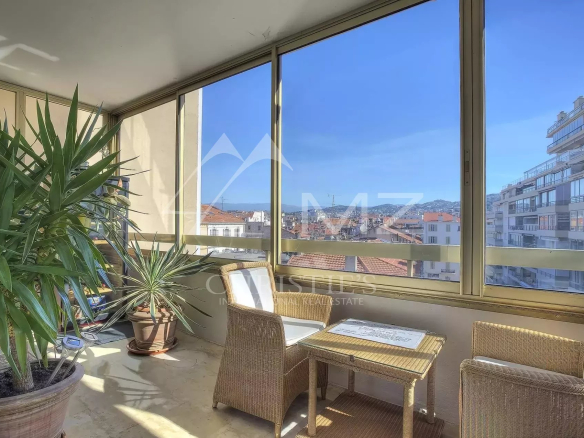 Sunlit enclosed balcony with wicker chairs and a small table, potted plants, and a cityscape view of red rooftops and modern buildings.