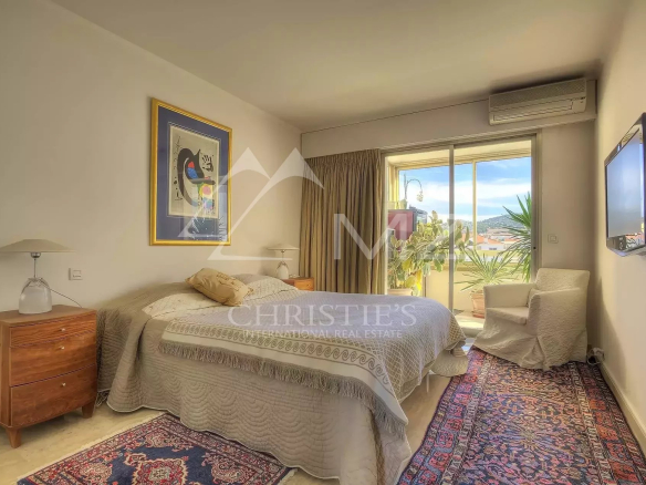 Sunlit hotel bedroom with a queen bed, two wooden nightstands, lamps, and a sliding glass door to a balcony with potted plants.