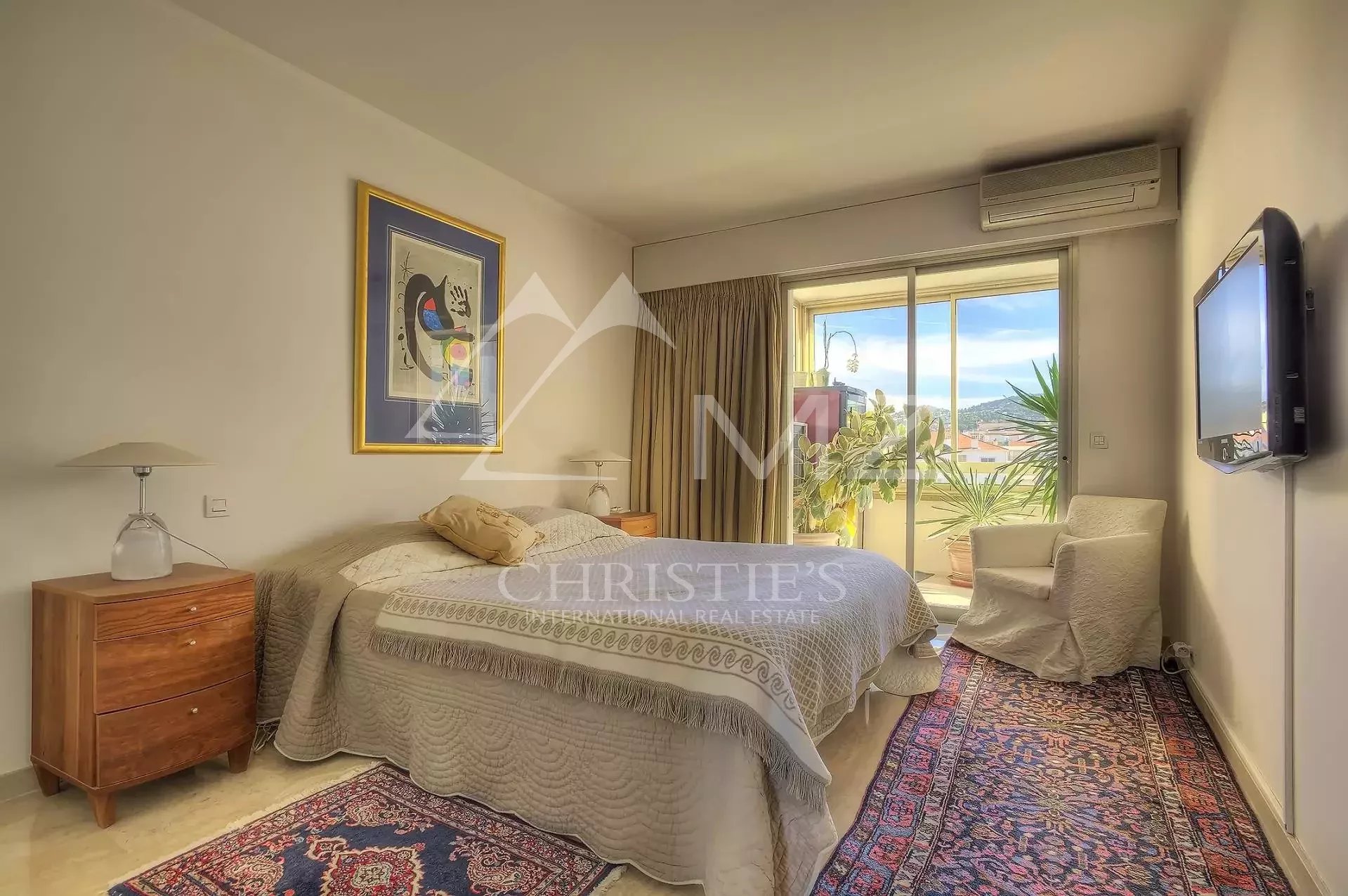 Sunlit hotel bedroom with a queen bed, two wooden nightstands, lamps, and a sliding glass door to a balcony with potted plants.