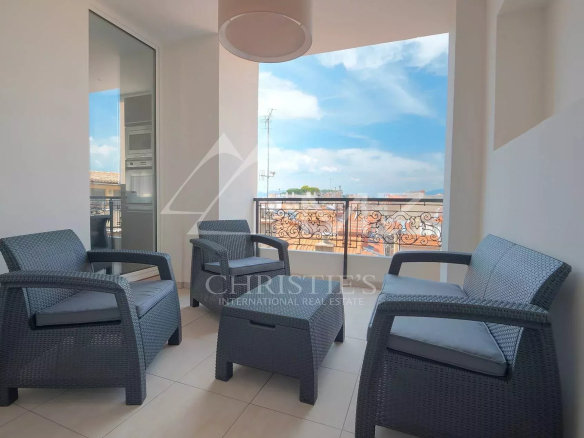Gray wicker patio seating on a balcony with four chairs around a matching table, cushions on the seats, overlooking a city skyline and blue sky.