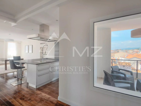 Bright open-plan kitchen with a stainless steel island and two bar stools, adjacent dining area, and a balcony overlooking city rooftops.