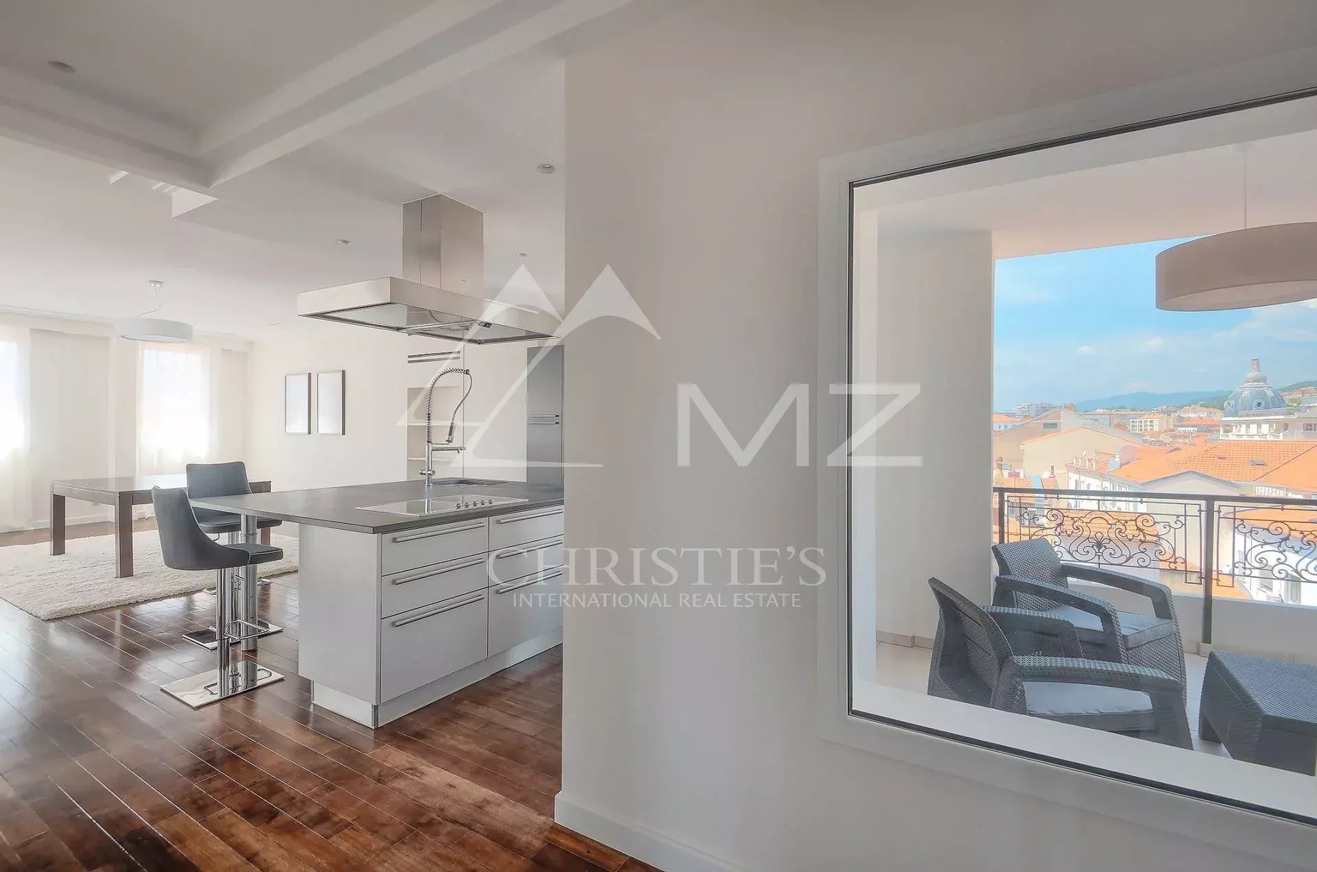Bright open-plan kitchen with a stainless steel island and two bar stools, adjacent dining area, and a balcony overlooking city rooftops.