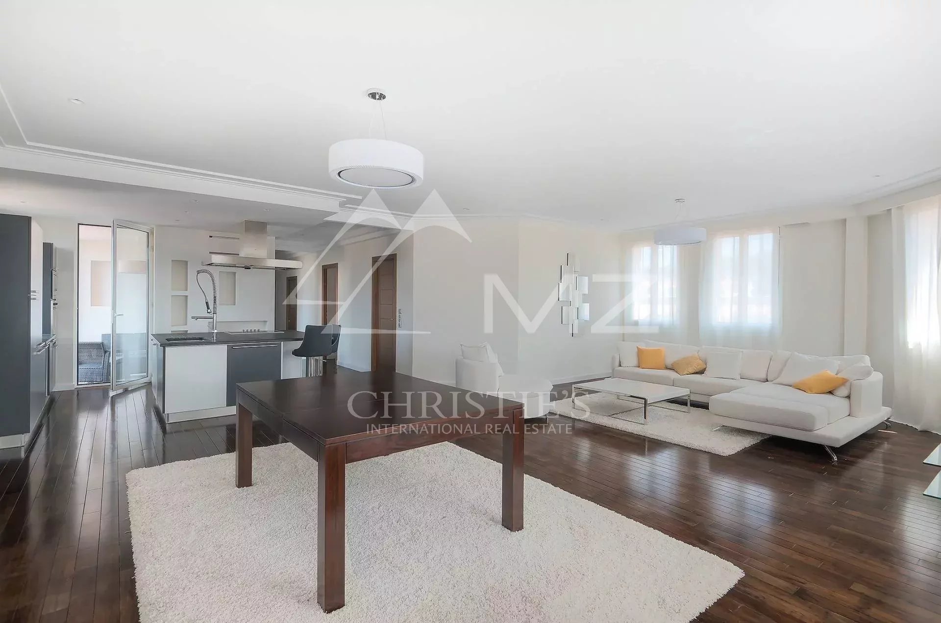Open-plan living area with a white sectional sofa, dark wood dining table, and a modern kitchen; large windows brighten the space.