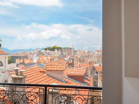 View over terracotta-tiled rooftops from a balcony, with a decorative wrought-iron railing in the foreground and a bright blue sky above a Mediterranean cityscape.