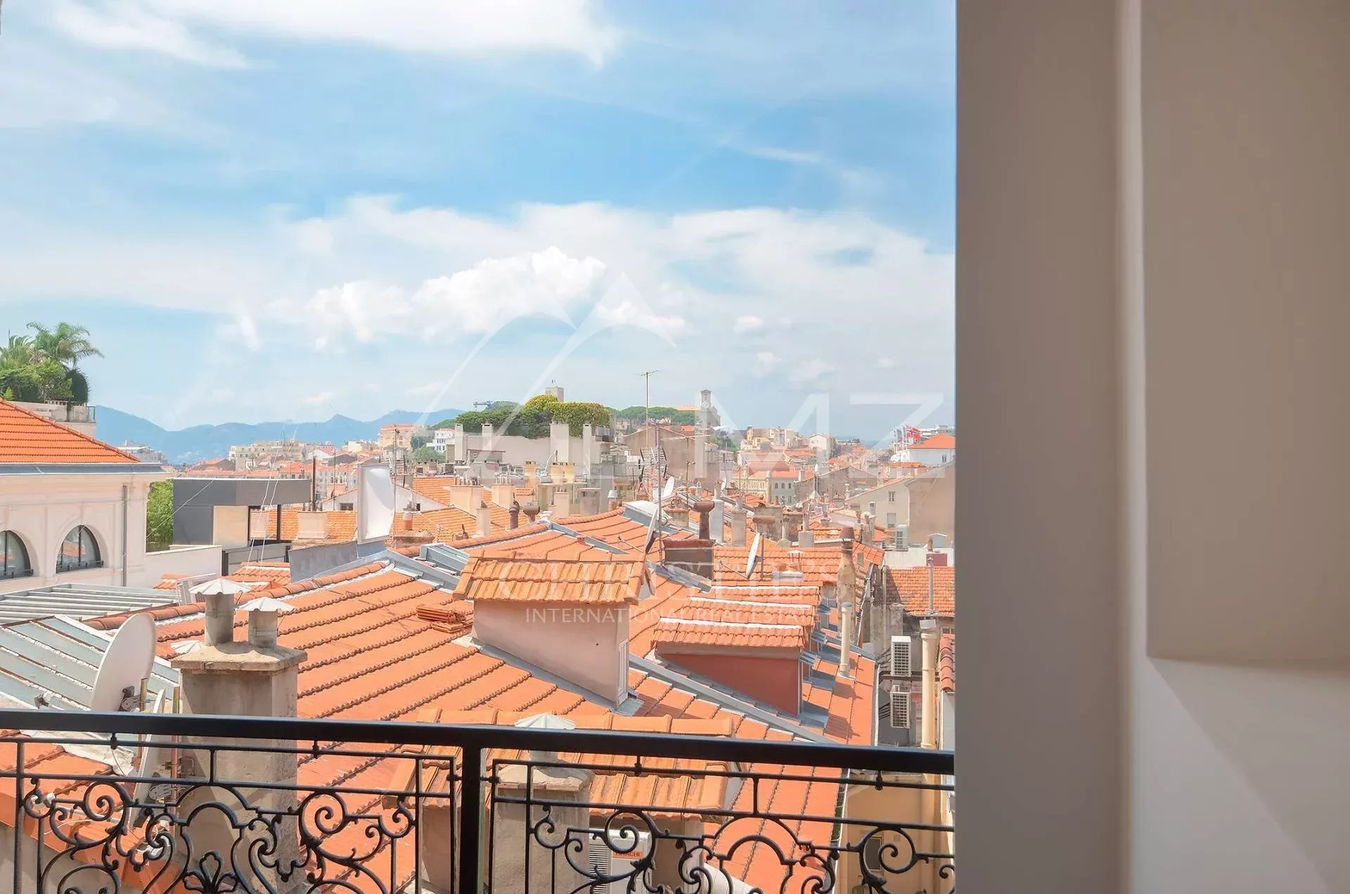 View over terracotta-tiled rooftops from a balcony, with a decorative wrought-iron railing in the foreground and a bright blue sky above a Mediterranean cityscape.