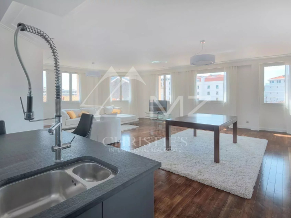 Open-plan living area with white sofa, large windows, and a kitchen island featuring a stainless steel sink in the foreground.