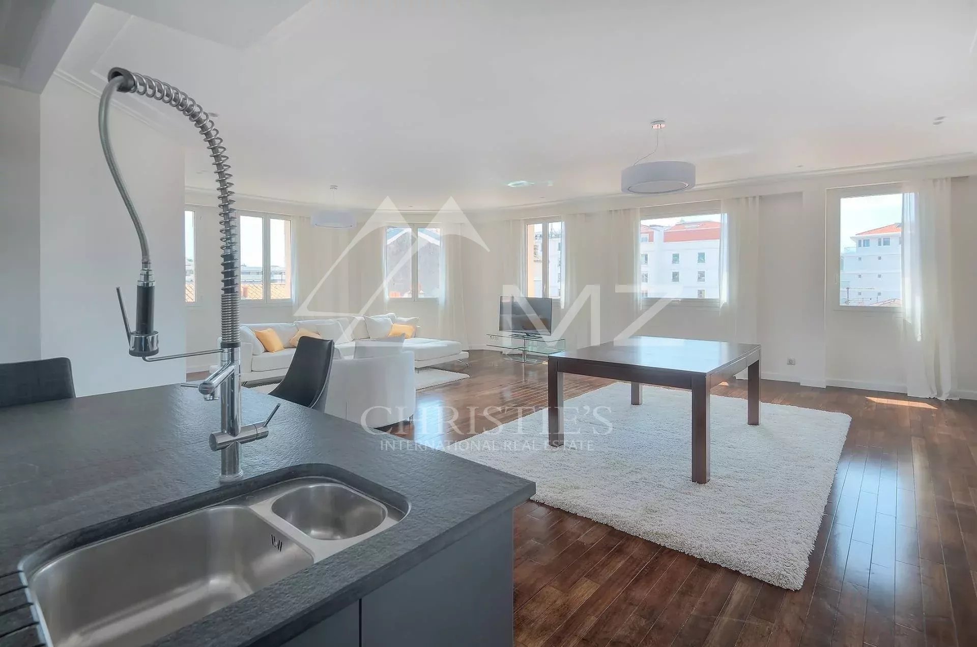 Open-plan living area with white sofa, large windows, and a kitchen island featuring a stainless steel sink in the foreground.