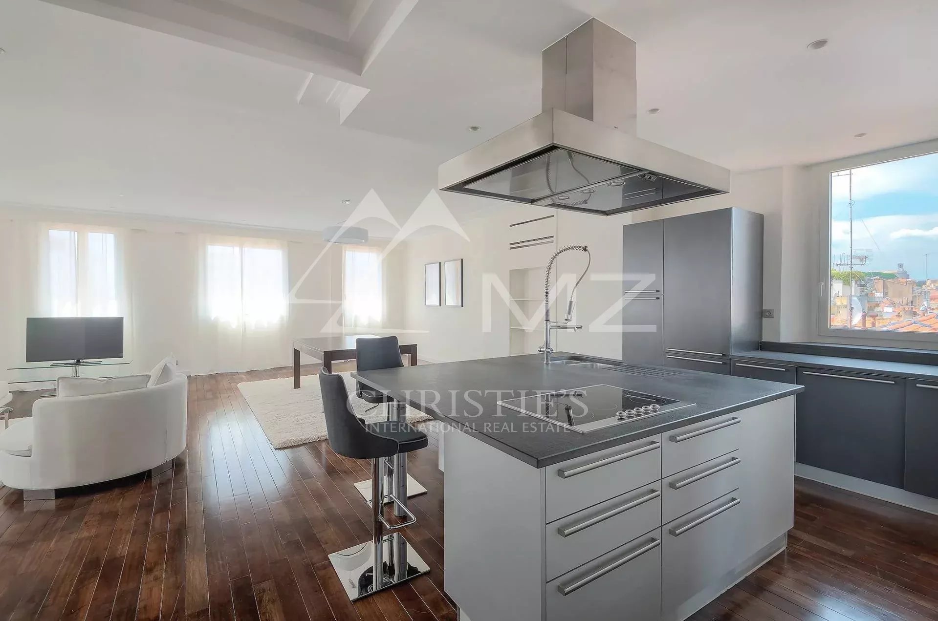 Open-concept kitchen with a stainless steel island, built-in cooktop and large range hood, dark cabinets, and a living area with white seating nearby.
