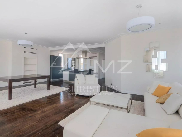 Open-plan living room with white sofas, glass coffee table, dark wood floors, and blue kitchen cabinetry in the background, showing an airy space.