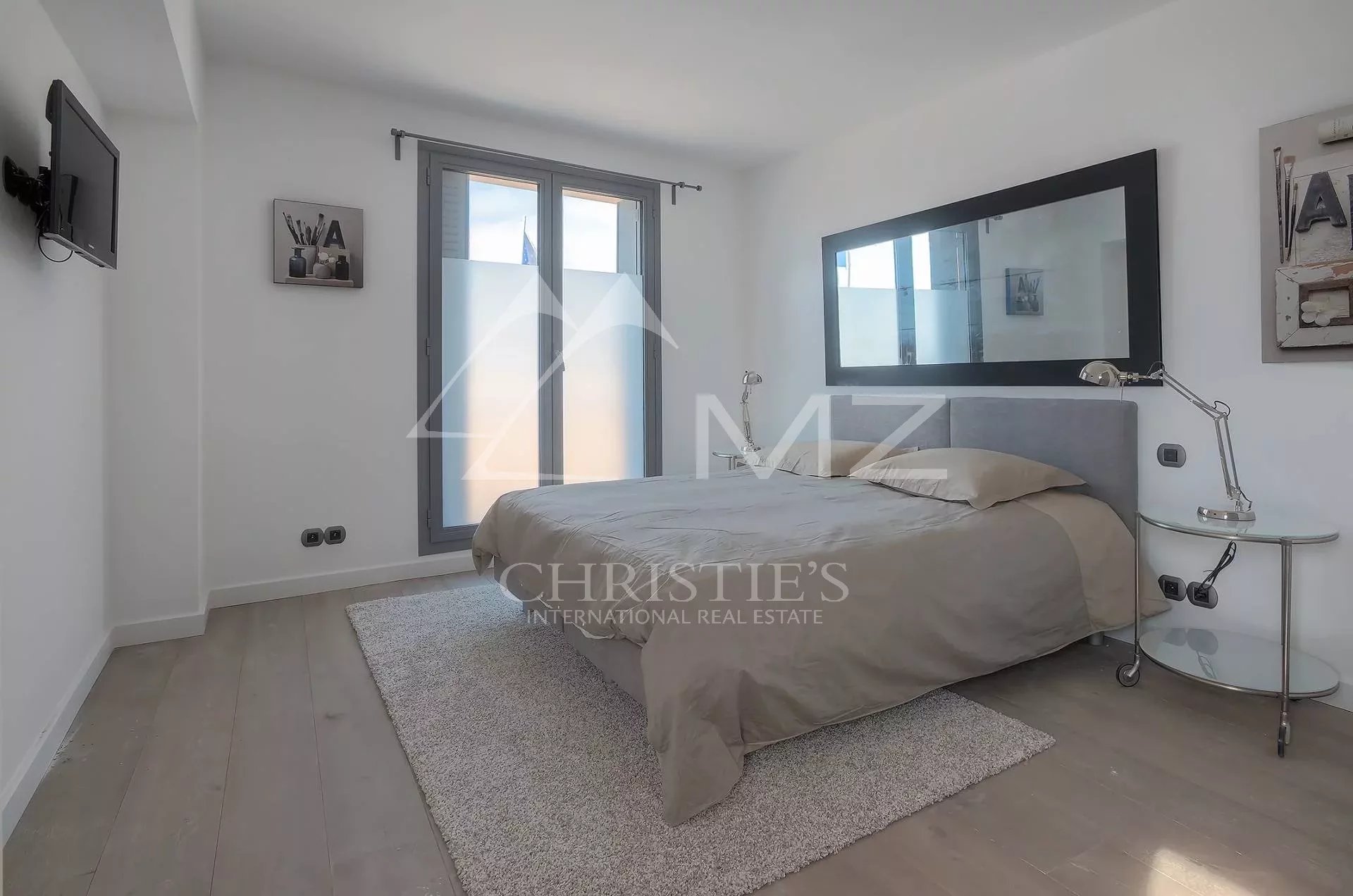 Bright minimalist bedroom with a gray upholstered bed, large black-framed mirror, and glass side table lamp on the right.