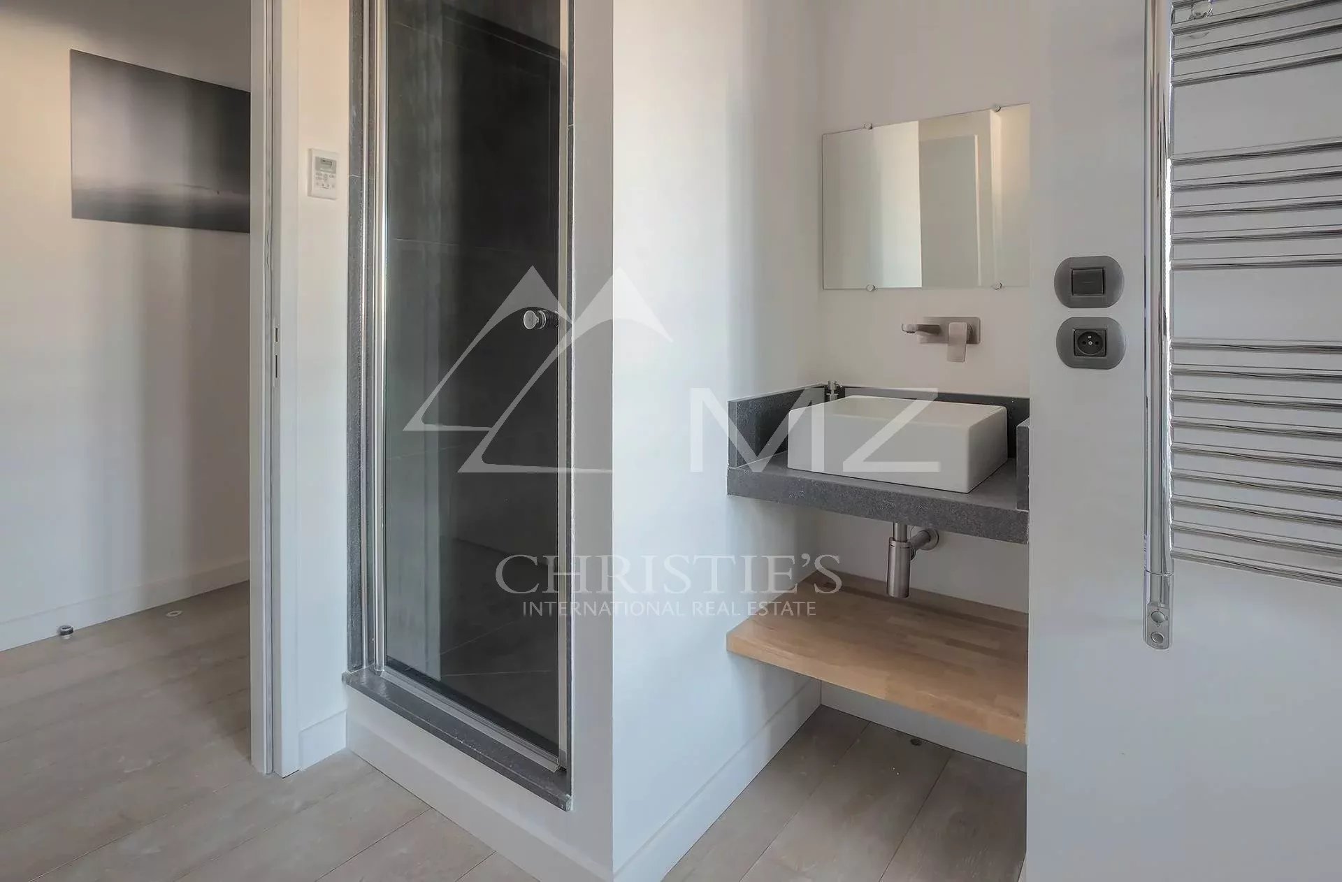 Modern bathroom with wall-mounted rectangular sink on a gray countertop, a wood shelf beneath, a mirror above, and a chrome towel warmer on the right; glass shower enclosure on the left.