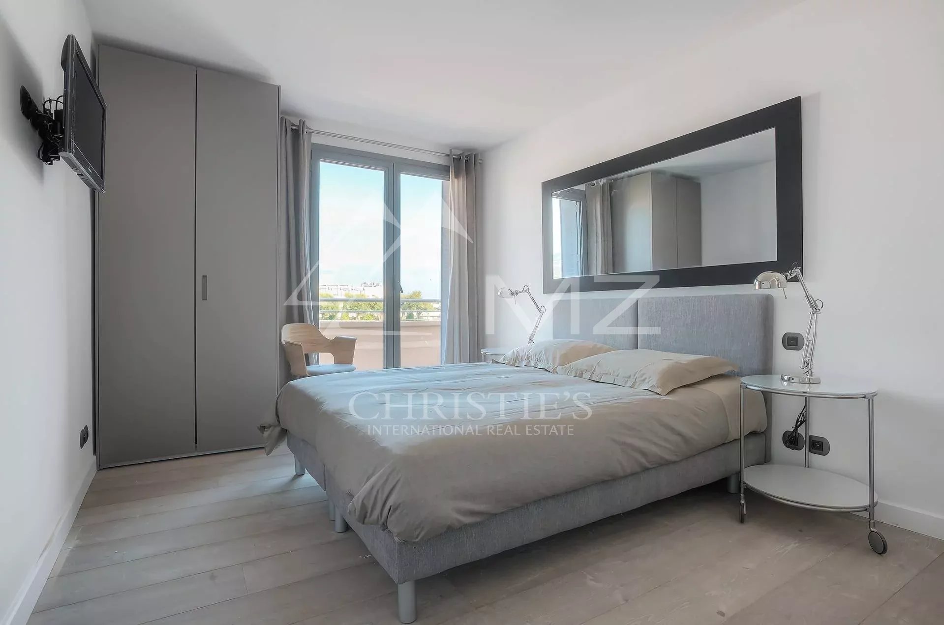 Modern bedroom with a gray upholstered bed, mirrored headboard, chrome bedside tables, a large framed mirror, and a wardrobe near a sliding balcony door.