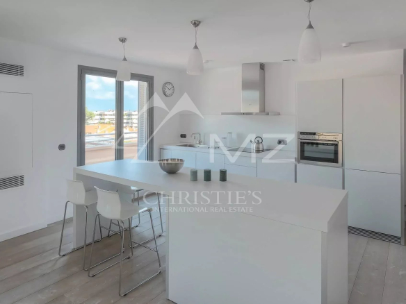 Bright white kitchen with a large central island, white bar stools, pendant lights, and a balcony door revealing an urban view.